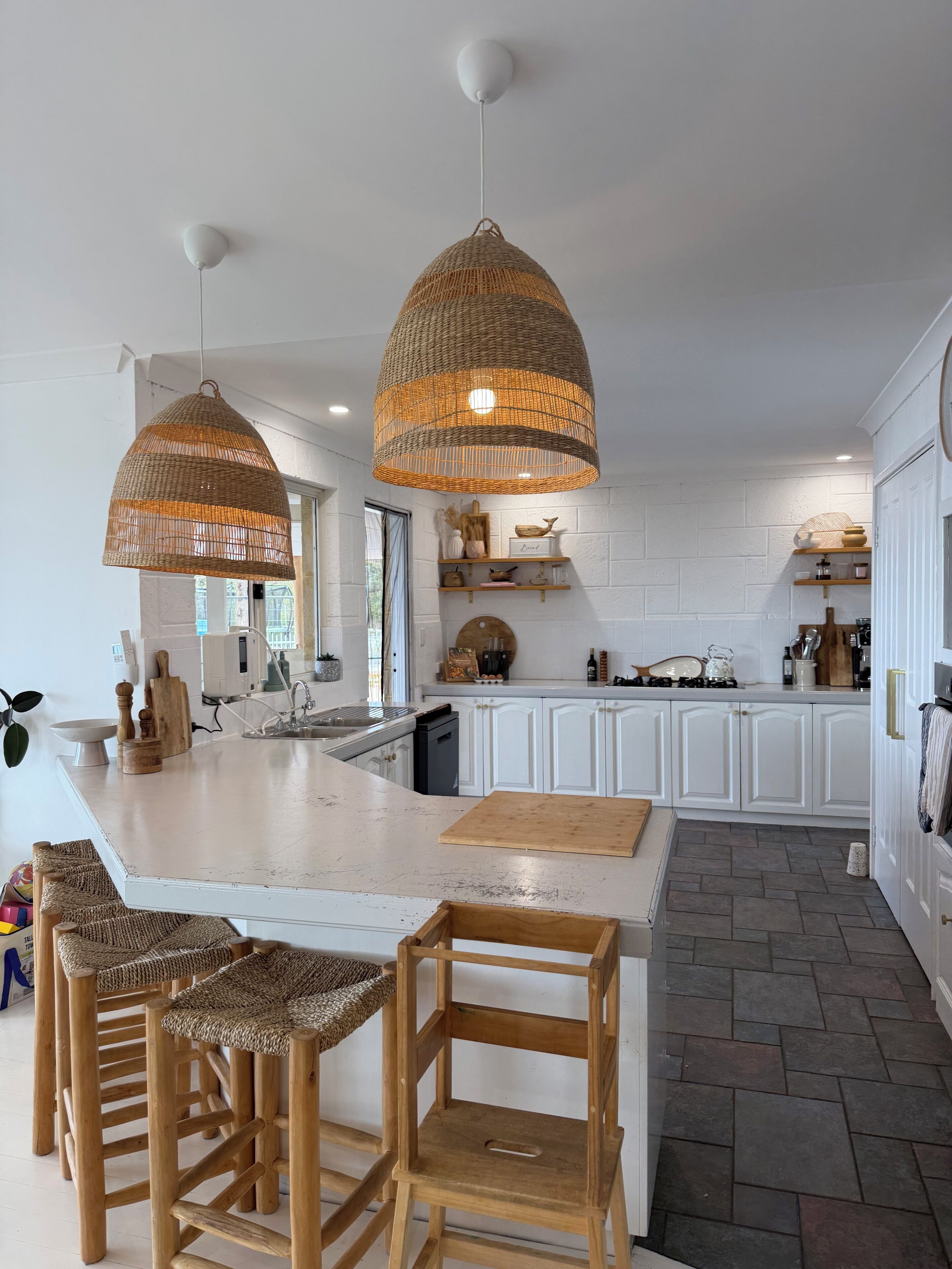 Une cuisine dotée d’un grand îlot central, de deux suspensions en osier, de meubles blancs et d’un sol carrelé à motifs en chevrons.