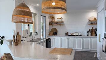 Une cuisine dotée d’un grand îlot central, de deux suspensions en osier, de meubles blancs et d’un sol carrelé à motifs en chevrons.