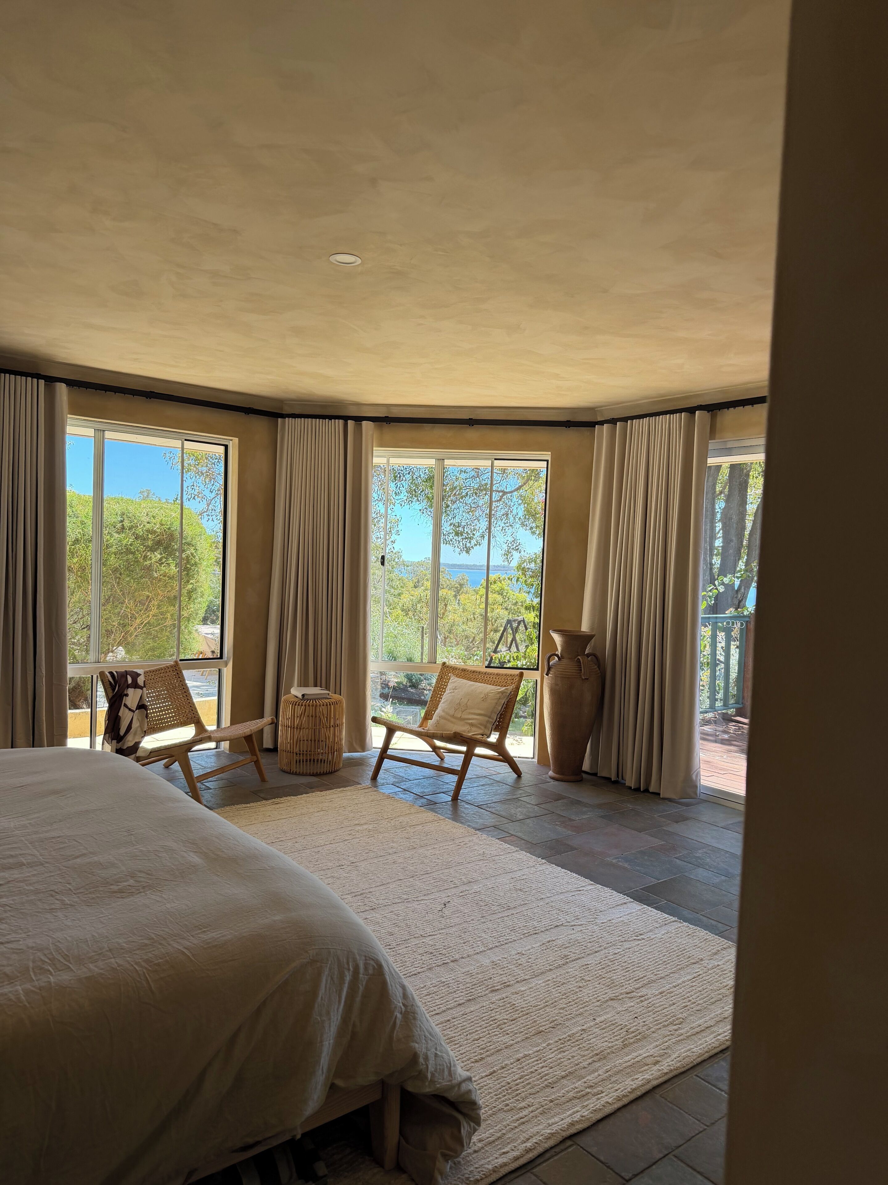 A bedroom with a large bed, two wicker chairs, a large vase, and a view of greenery and a body of water through large windows.