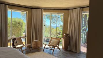 A bedroom with a large bed, two wicker chairs, a large vase, and a view of greenery and a body of water through large windows.