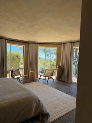Ein Schlafzimmer mit einem großen Bett, zwei Korbstühlen, einer großen Vase und Blick auf Grünflächen und ein Gewässer durch große Fenster.