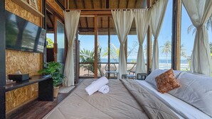 A room with a bed, a TV, a plant, and a view of the beach. - Salty Cabin Sunset Views Yeh Leh Beach (Selemadeg)