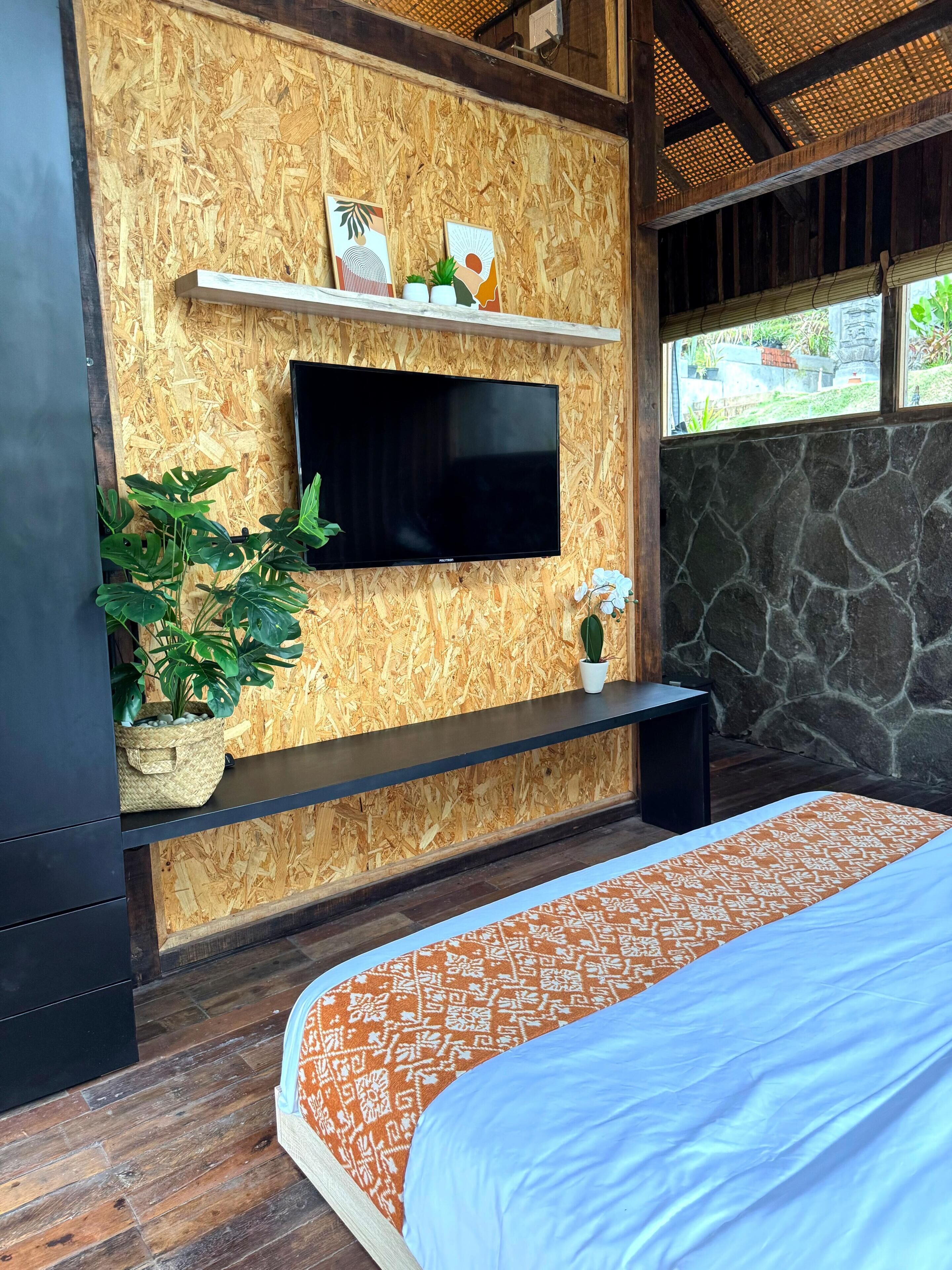 Une chambre à coucher comprenant un lit, un téléviseur fixé au mur, une étagère avec des objets décoratifs et une plante en pot.