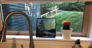 A window with a metal faucet, a framed landscape painting, and a potted cactus.