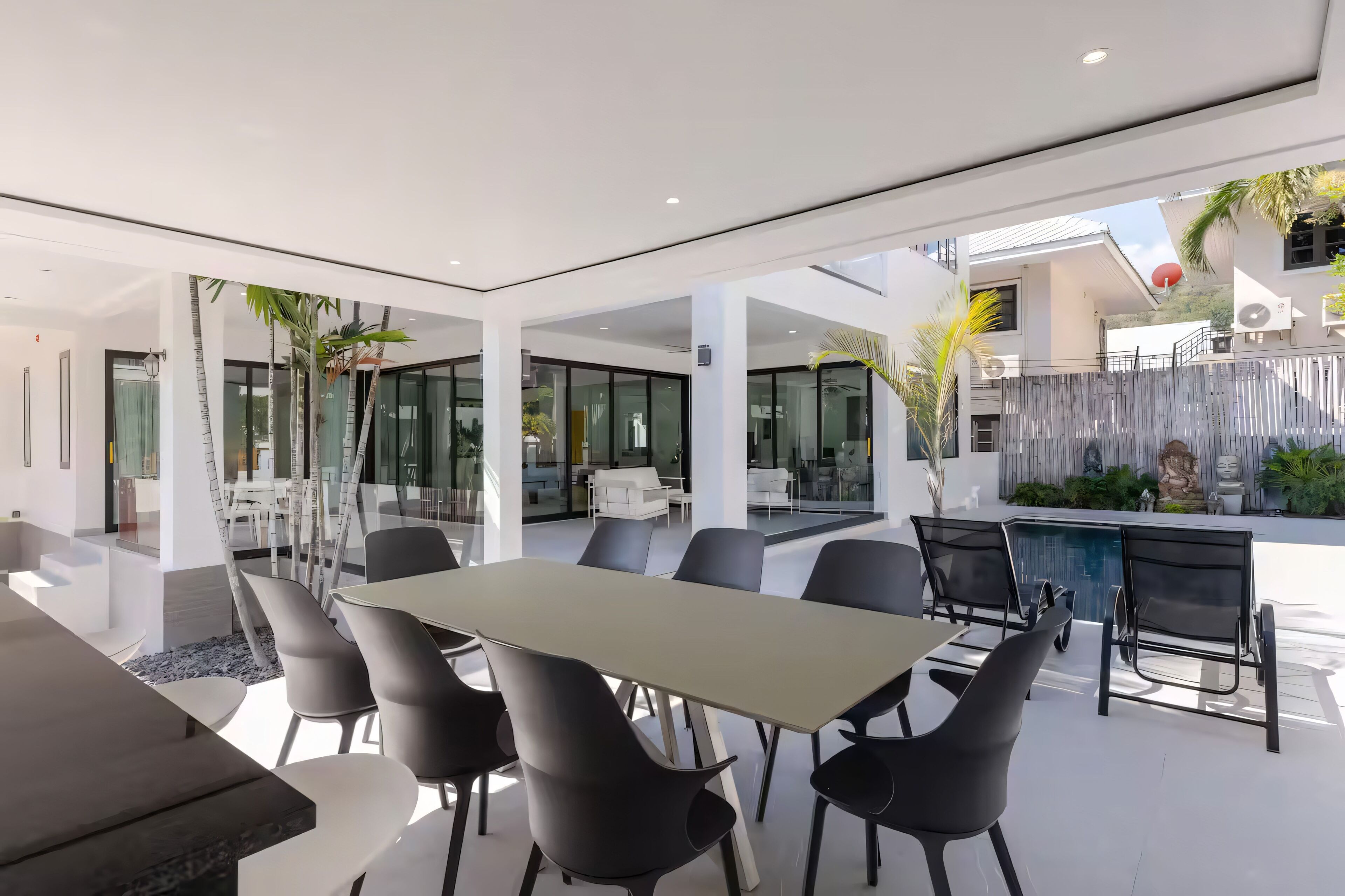 A modern outdoor dining area with a long table and chairs, surrounded by glass walls and a pool.
