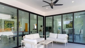 A modern interior with glass walls, white furniture, ceiling fans, and a dining area visible through the glass. - Grand Hill Mansion (Hua Hin)