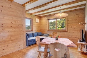 Interior de una cabaña de madera con una mesa de comedor, sillas, un sofá, un televisor y una ventana con vistas a la vegetación.