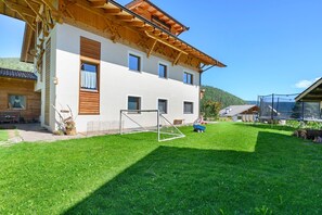 A two-story building with a wooden roof, a visible window, and a grassy yard with a goalpost.