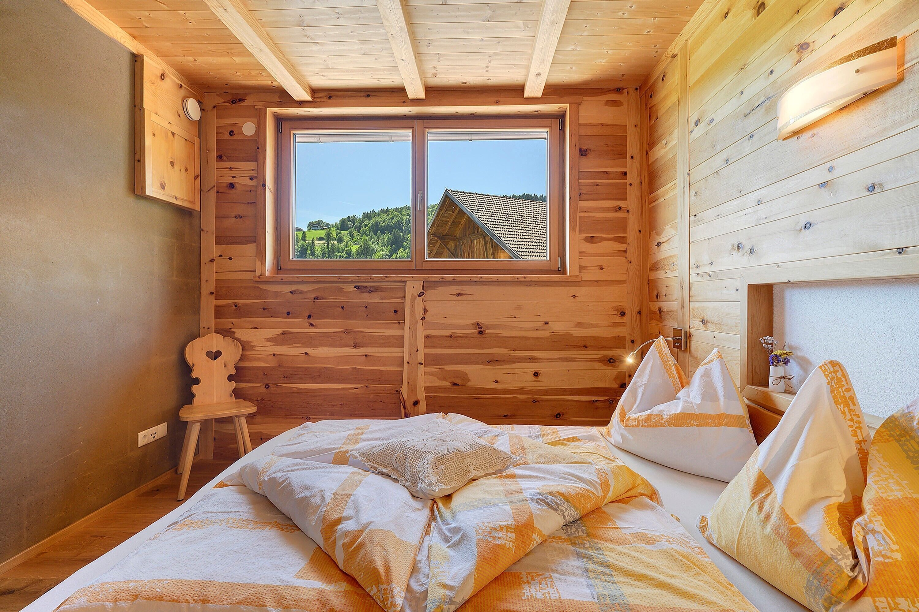 Een slaapkamer in een houten chalet, met een bed, een stoel en een raam met uitzicht op een huis.