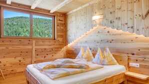 Un dormitorio de cabaña de madera con cama, almohadas, mesita de noche, ventana y una lámpara fijada a la pared.