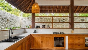 A kitchen with a stone wall, wooden cabinets, a sink, and a built-in oven. - Villa Calypso - 3 Br (Seminyak)