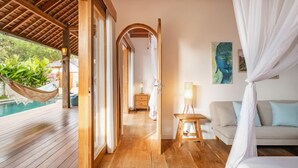 A room with a wooden floor, a bed with a canopy, a sofa, a small table, and a painting on the wall. - Villa Calypso - 3 Br (Seminyak)