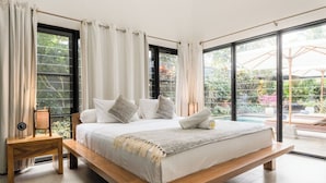 Ein großes Bett mit hölzernem Gestell, weißer Bettwäsche und Blick auf einen Garten durch große Fenster.