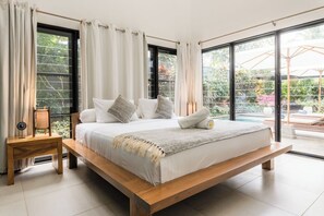 Ein großes Bett mit hölzernem Gestell, weißer Bettwäsche und Blick auf einen Garten durch große Fenster.