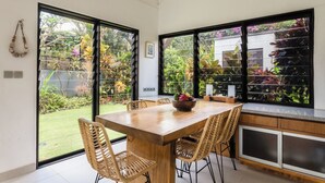 Ein Essbereich mit Holztisch und Korbstühlen, große Fenster mit Gartenblick und eine Küchenzeile mit Schränken und Arbeitsplatte.