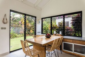 Ein Essbereich mit Holztisch und Korbstühlen, große Fenster mit Gartenblick und eine Küchenzeile mit Schränken und Arbeitsplatte.