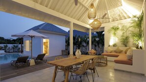 An outdoor dining area with a wooden table and chairs, a sofa, and a pool in the background. - Bingin Beach Villas (Pecatu)