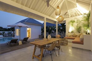 An outdoor dining area with a wooden table and chairs, a sofa, and a pool in the background. - Bingin Beach Villas (Pecatu)
