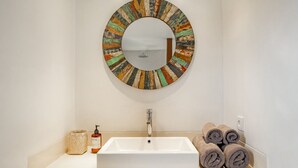 Une salle de bain avec un lavabo blanc, un miroir rond doté d’un cadre coloré, un distributeur de savon, un porte-serviettes avec des serviettes pliées, et un petit panier en osier.