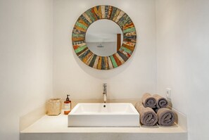 A bathroom with a white sink, a round mirror with a colorful frame, a soap dispenser, a towel holder with rolled towels, and a small woven basket. - Batubolong - Studio (Canggu)