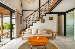 A modern living room with a sofa, coffee table, staircase, and a view through a glass door. - Batubolong - Studio (Canggu)