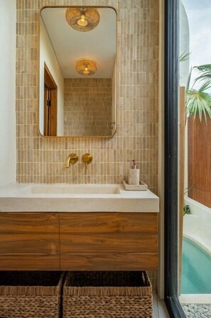 Ein Badezimmer mit Fliesen, einem Spiegel, einem Waschbecken mit zwei Wasserhähnen und einem Fenster mit Blick auf einen Pool.