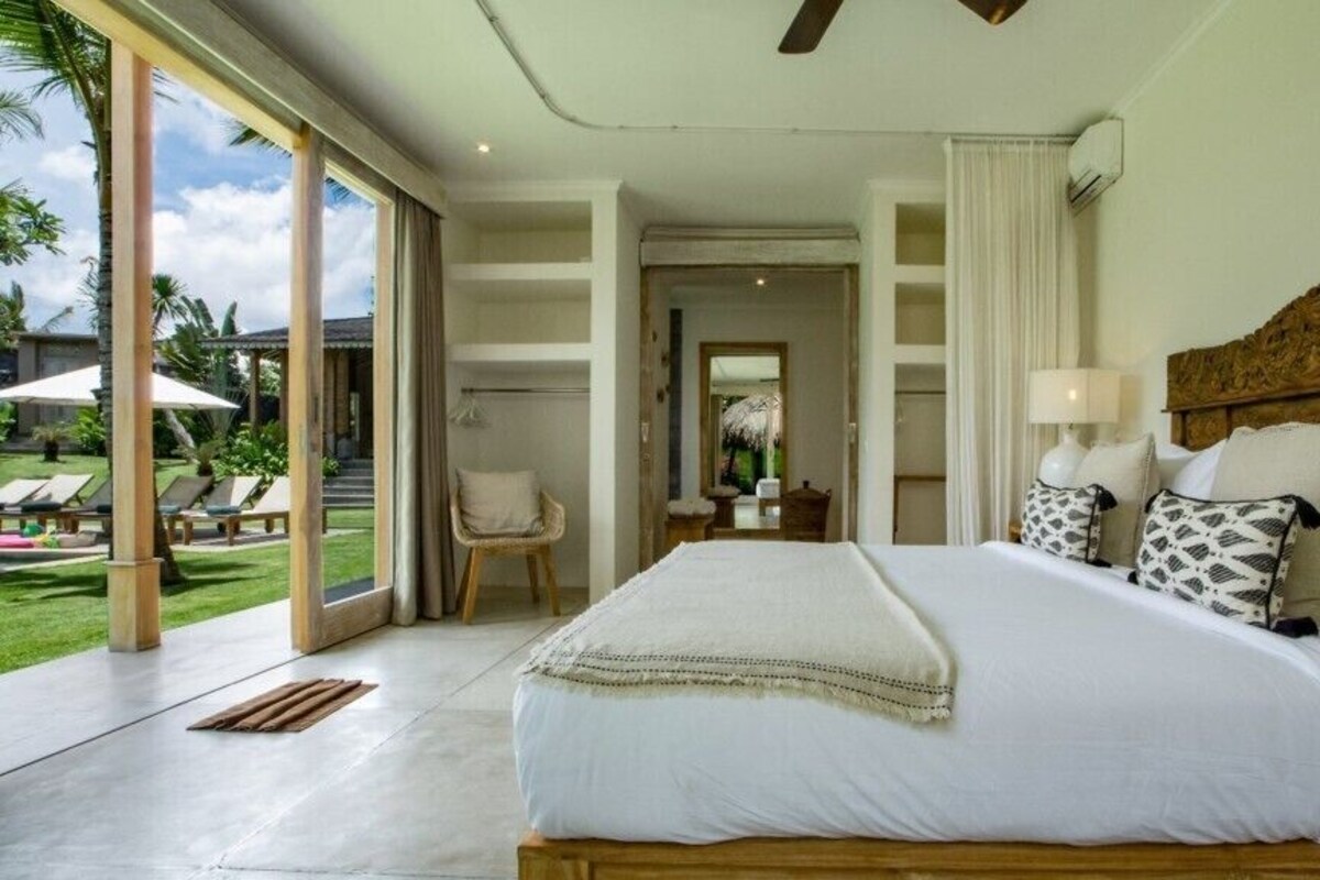 A spacious bedroom with a large bed, a wooden headboard, and a view of a lush garden through large sliding doors.