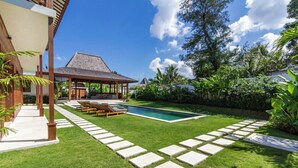 Un patio trasero con piscina, una zona de estar techada y un césped bien cuidado.