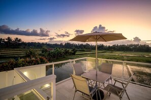 A terrace with a table, chairs, and an umbrella overlooking a rice field at sunset. - Villa Chai - 2 Br (Ubud)