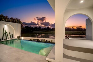 A modern pool area with a view of a sunset over a field, a white infinity pool, and a curved seating area. - Villa Chai - 2 Br (Ubud)