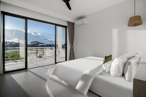 A modern hotel room with a large sliding glass door leading to a balcony, a bed with white linens, and a ceiling fan. - Villa Chai - 2 Br (Ubud)