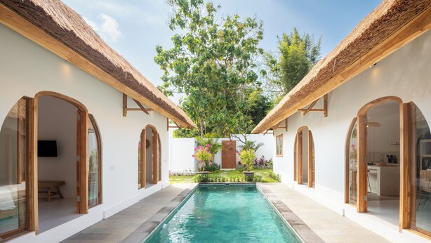 Una piscina de agua cristalina azul, rodeada de muros blancos y arcos de madera, con un árbol y flores al fondo.