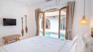 Un dormitorio con una cama grande, televisor, unas palas de madera, un banco y vista a una piscina.