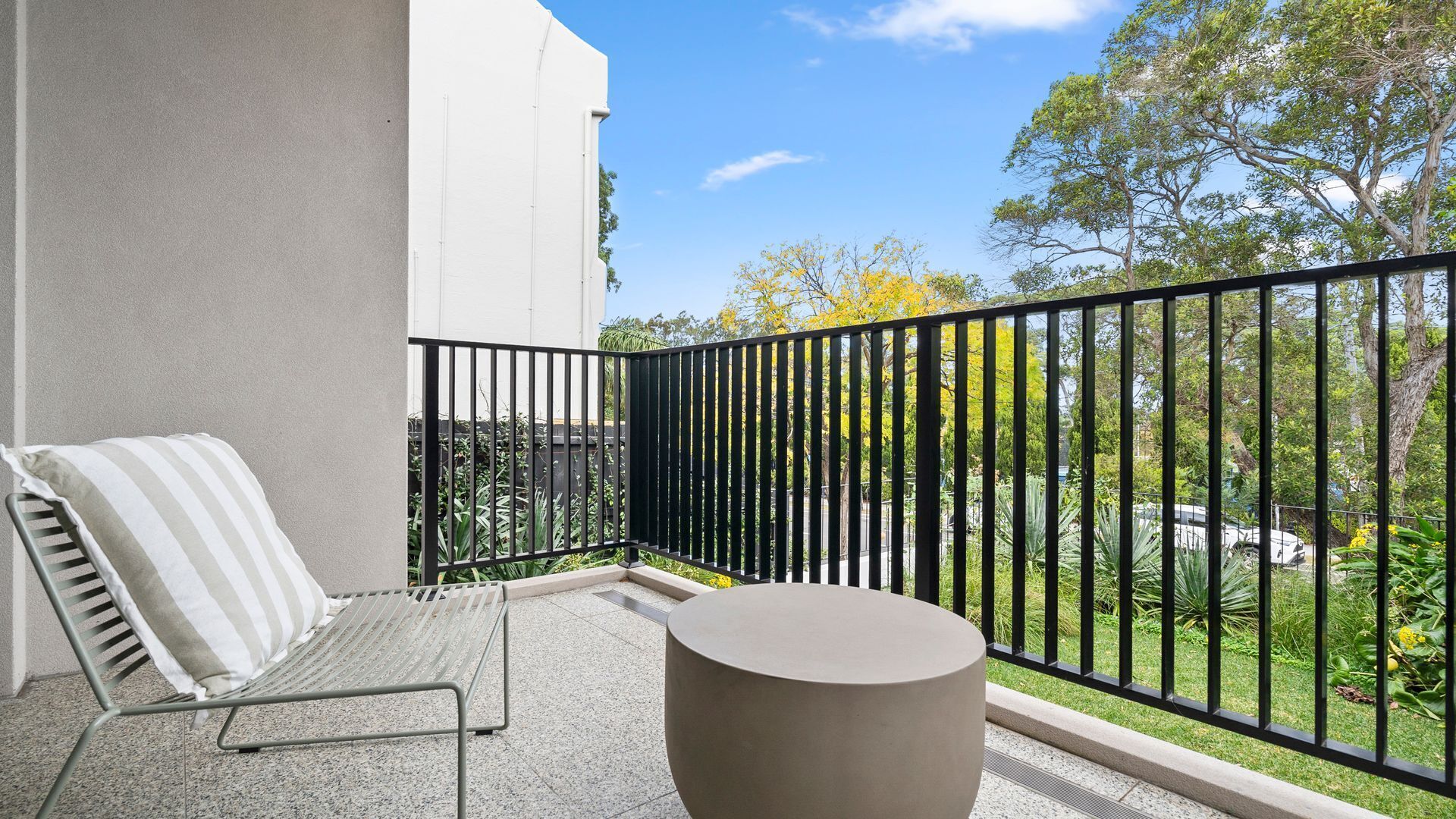 Un balcón con una silla metálica y una mesa redonda, una cerca de metal negro, y vistas a vegetación y un edificio.