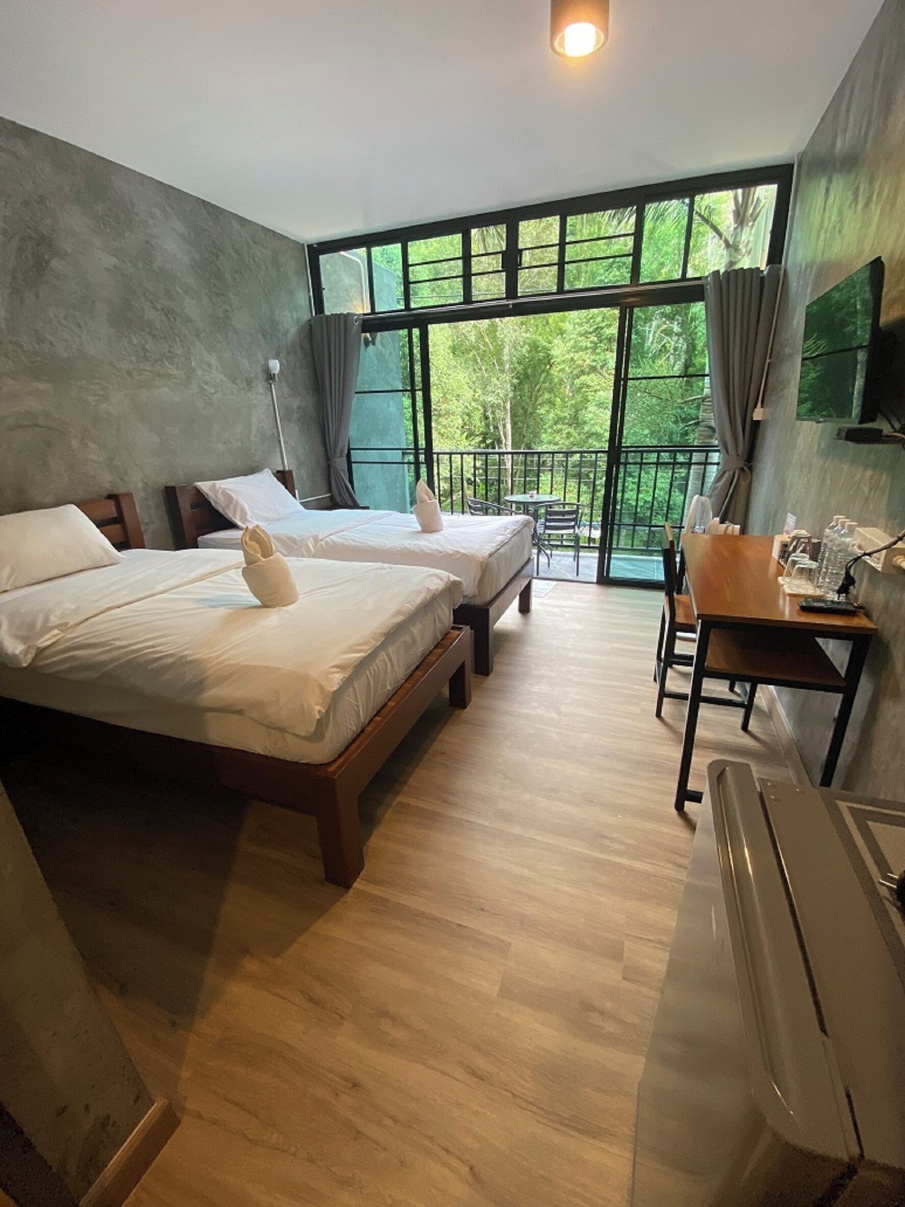 A hotel room with two beds, a desk, a chair, and a large window with a view of greenery.