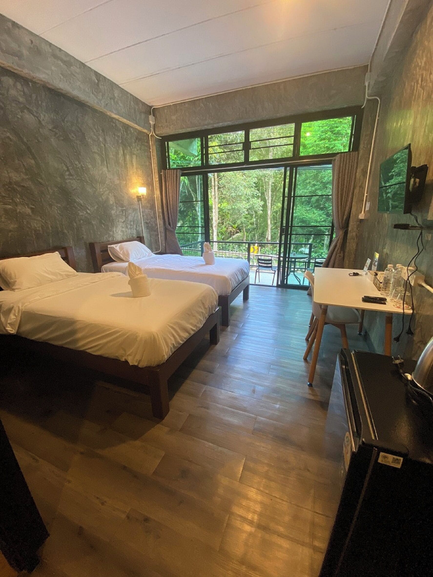 A hotel room with two beds, a dining table, and a view of greenery outside.
