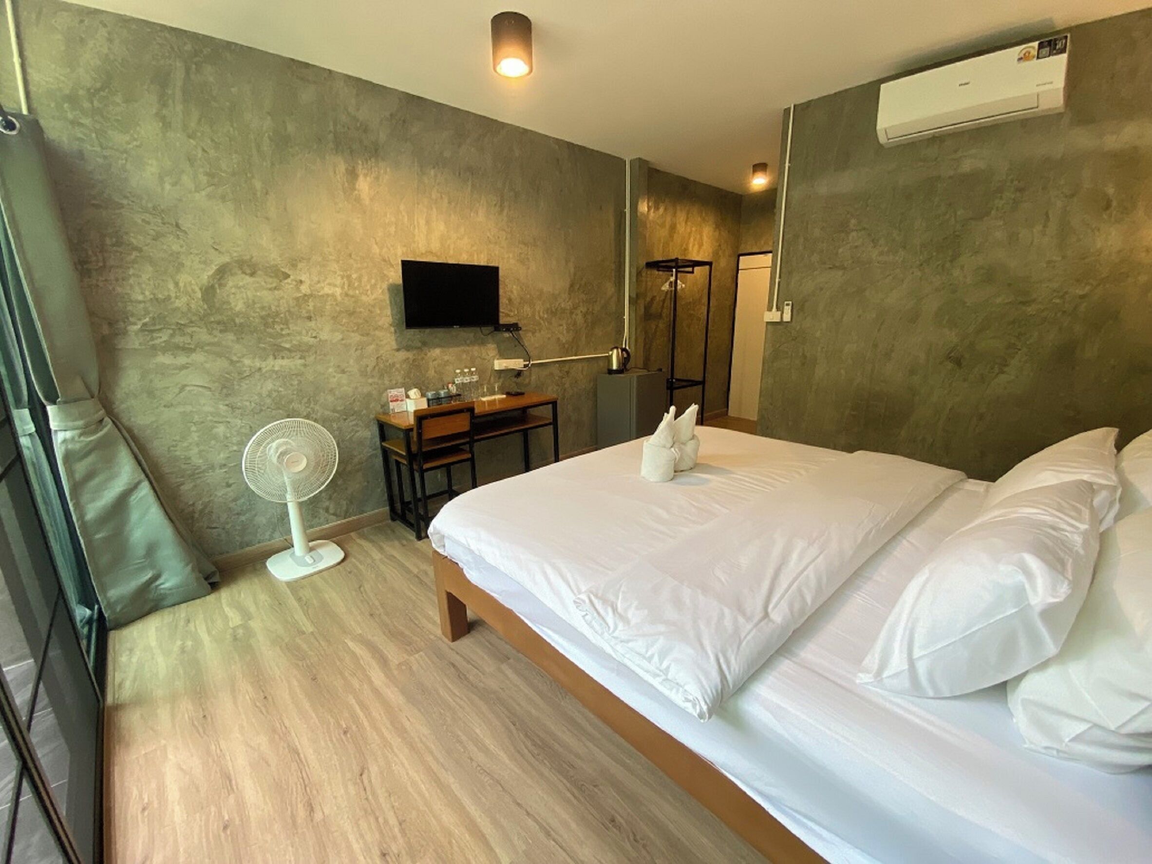 Habitación de hotel con cama, escritorio, silla, ventilador de pie, televisor montado y equipo de aire acondicionado.