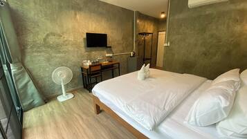 Habitación de hotel con cama, escritorio, silla, ventilador de pie, televisor montado y equipo de aire acondicionado.