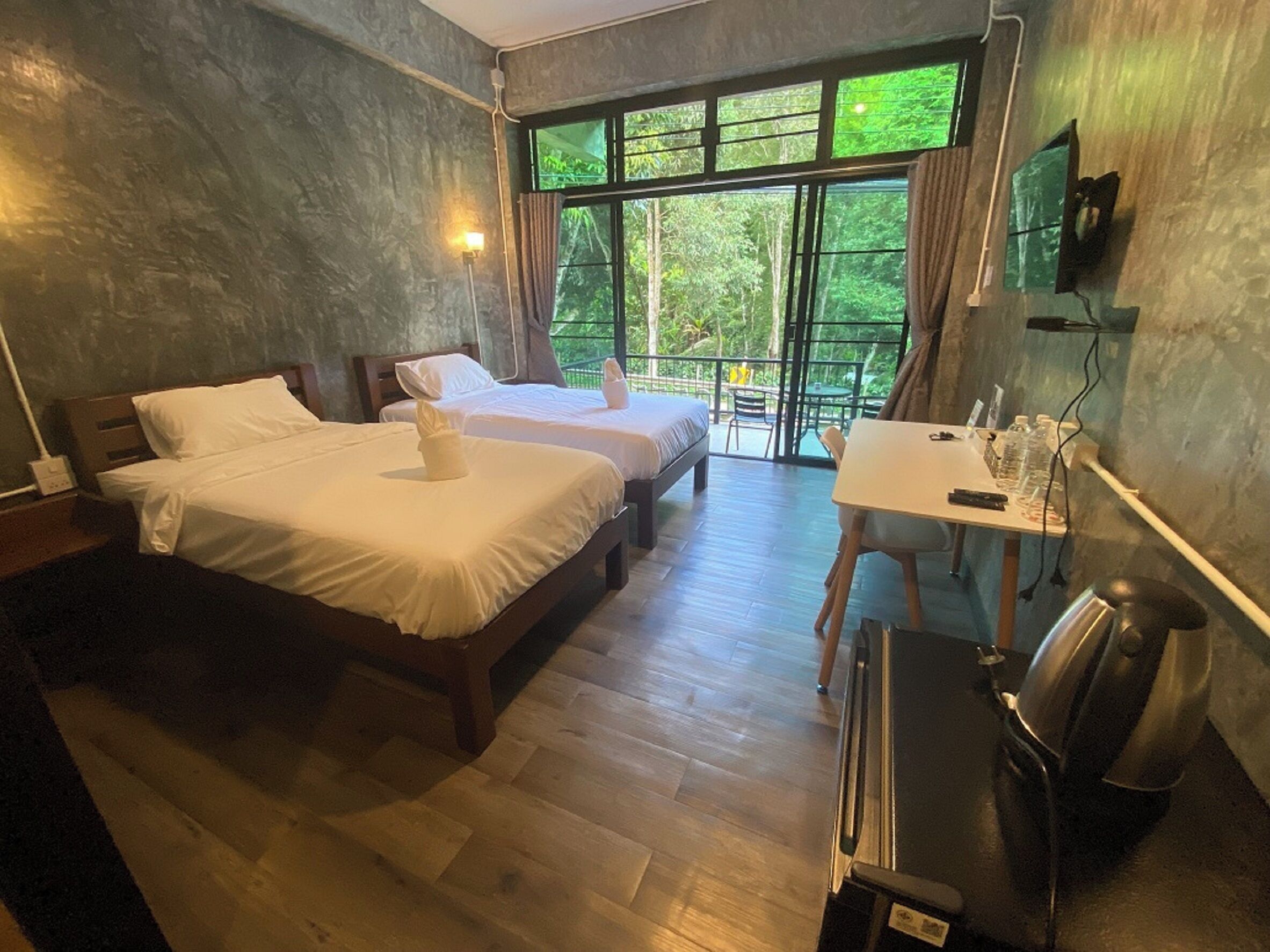 Een hotelkamer met twee bedden, een bureau, een stoel, een televisie en een balkon met uitzicht op groen.