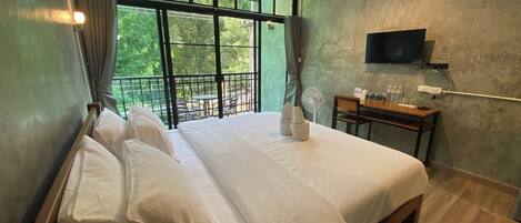 Un lit double avec du linge de lit blanc, une petite table avec un ventilateur, un téléviseur fixé au mur et un balcon donnant sur un paysage verdoyant.