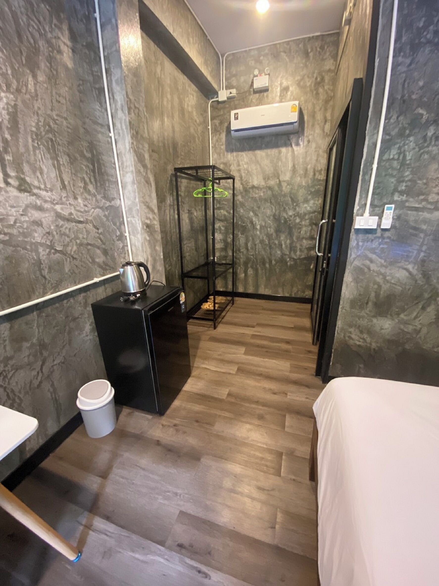Una habitación compacta con una cama, un escritorio pequeño con hervidor, un cubo de basura y una planta colgante.