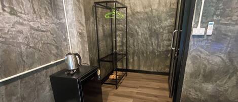 Una habitación compacta con una cama, un escritorio pequeño con hervidor, un cubo de basura y una planta colgante.