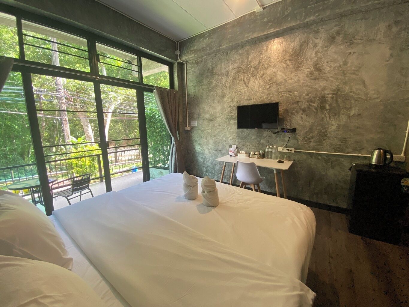 A hotel room with a large bed, a desk, a TV, and a view of greenery through the window.