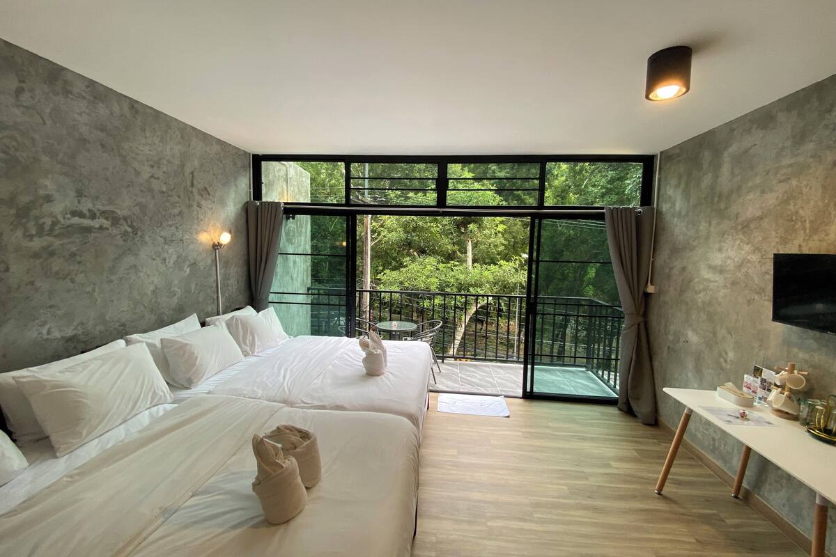 A modern hotel room with a large bed, a small table, a TV, and a balcony with a view of greenery.