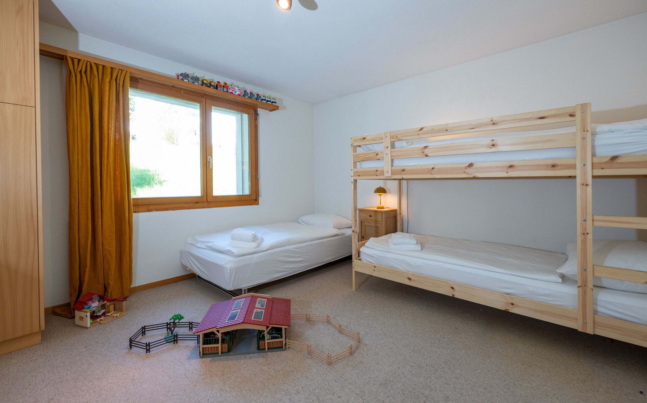Une chambre avec deux lits superposés, une fenêtre avec des rideaux, une petite table de chevet avec une lampe et une maison de jeu posée sur le sol.