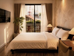 A hotel room with a bed, a nightstand, a lamp, a potted plant, and a view of a cityscape through a large window. - Amana Living Nguyen Canh Chan (Cau Ong Lanh)
