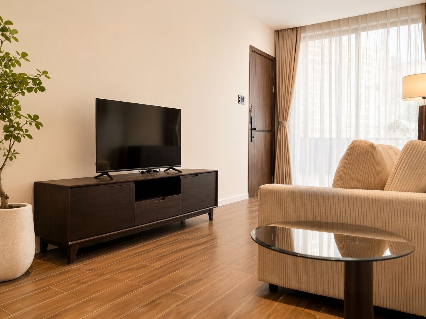 Ett modernt vardagsrum med en platt-TV placerad på ett mörkt träskåp, en beige soffa, ett soffbord i glas och en krukväxt.