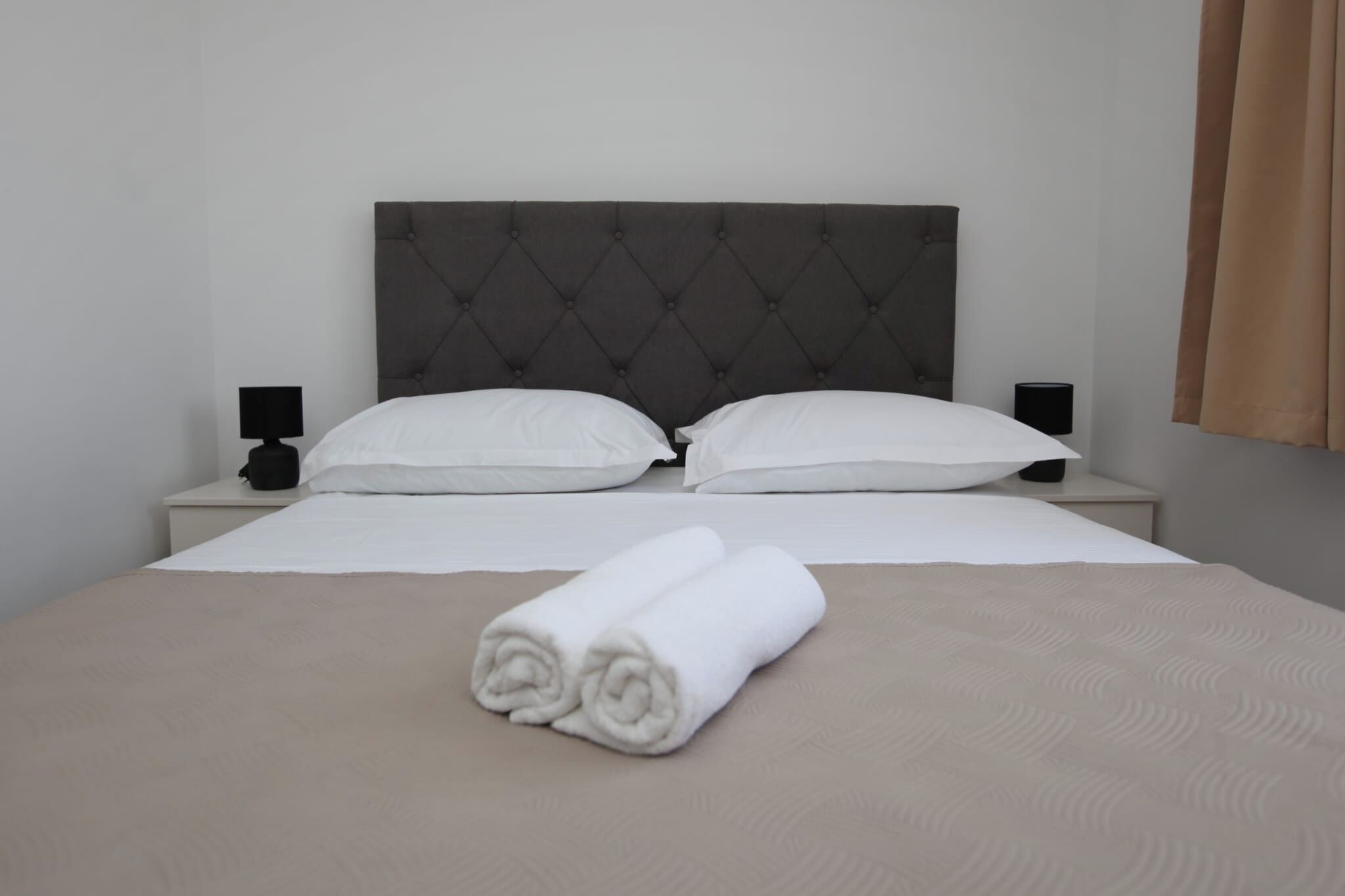 A neatly made bed with white linens, two pillows, and a rolled towel on the bed. There are two bedside tables with black lamps.