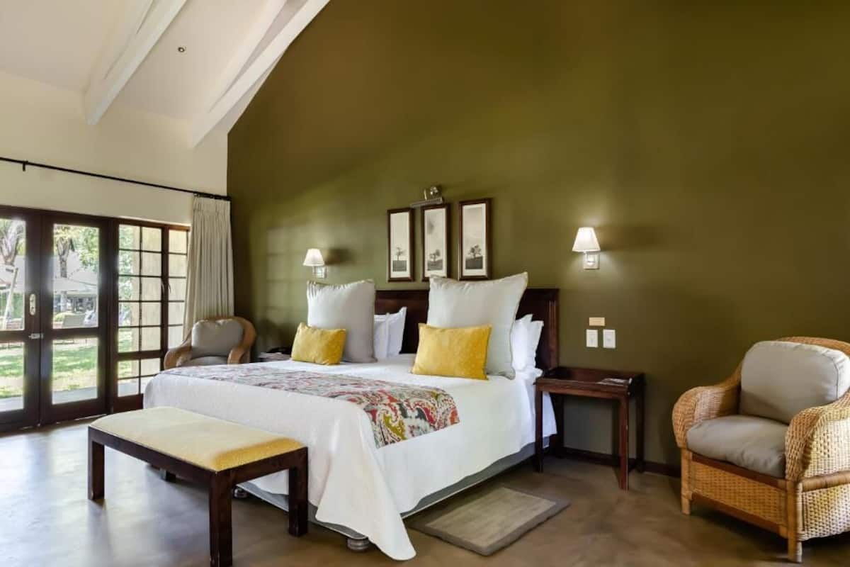 A bedroom with a large bed, a bench, a wicker chair, a nightstand, and a window with curtains.