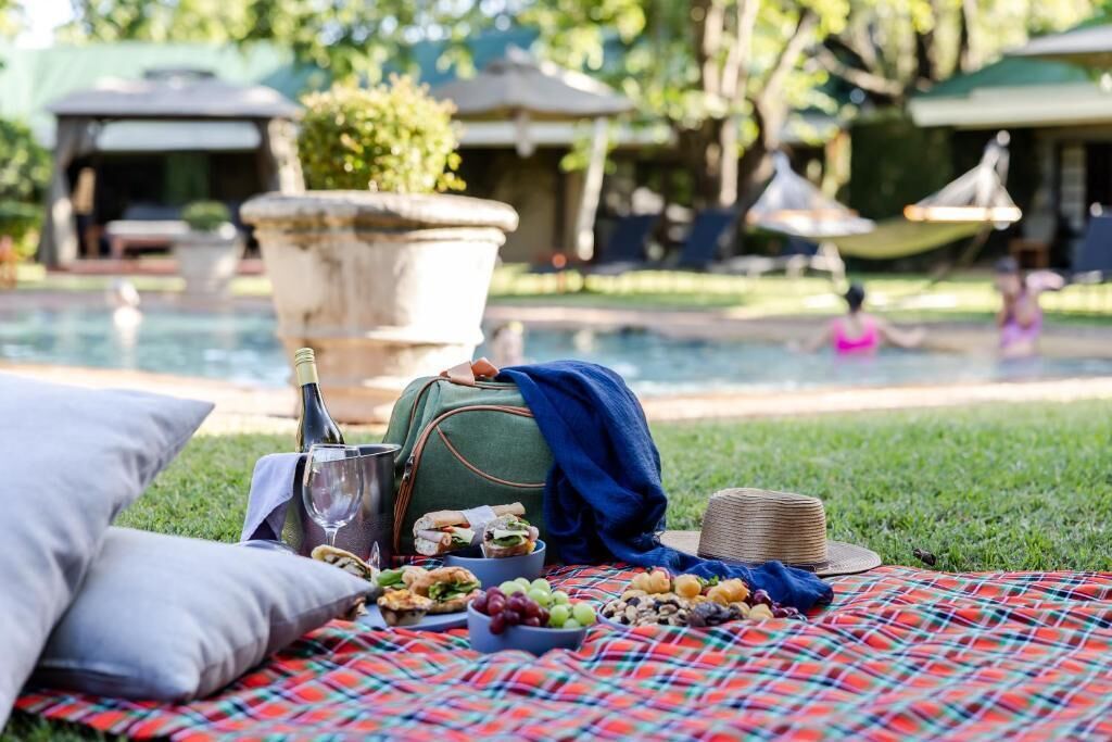 Un montaje para picnic con comida, una botella y un bolso sobre una manta junto a una piscina.